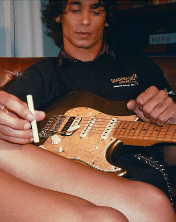 Close-up of a man on a leather couch holding a cigarette with a Strat-style electric guitar on his lap; another person’s legs in the foreground.