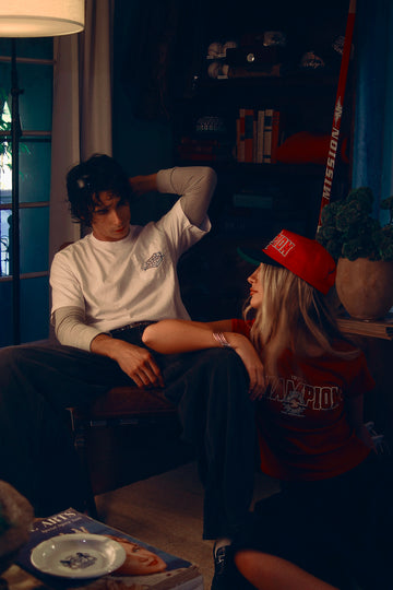 Moody indoor scene with a man in a white layered long-sleeve tee sitting on a chair and a woman in a red cap and T-shirt sitting on the floor, looking up at him
