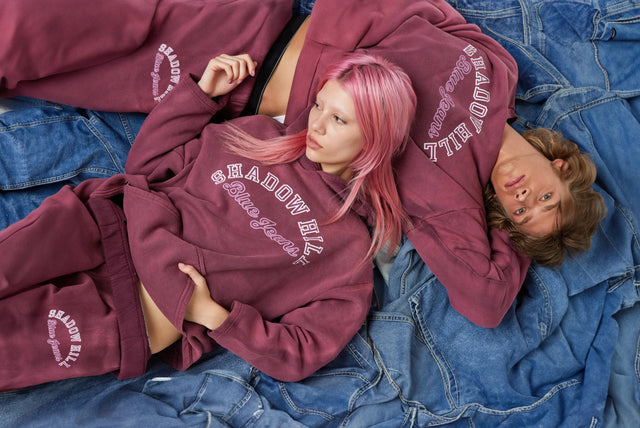 Two models lying on a bed of denim fabric, wearing matching maroon Shadow Hill sweatshirts and sweatpants with 'Blue Jeans' text.